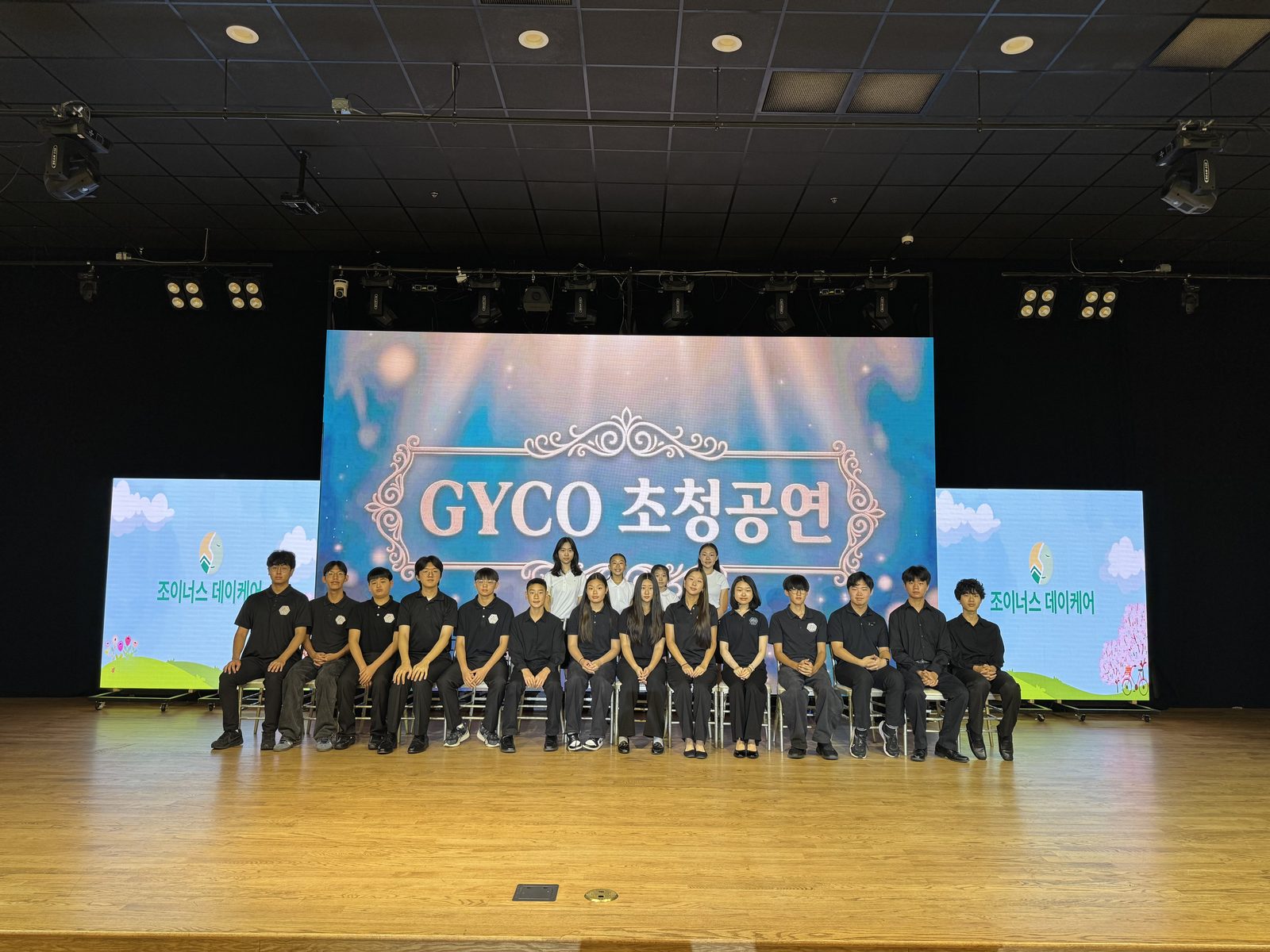 The GYCO ensemble seated on stage beneath the GYCO invited-performance screen at Joynus Daycare