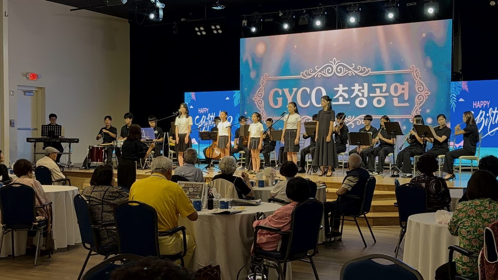 Young singers and the student orchestra performing for seniors seated at their tables during the Joynus Daycare birthday celebration
