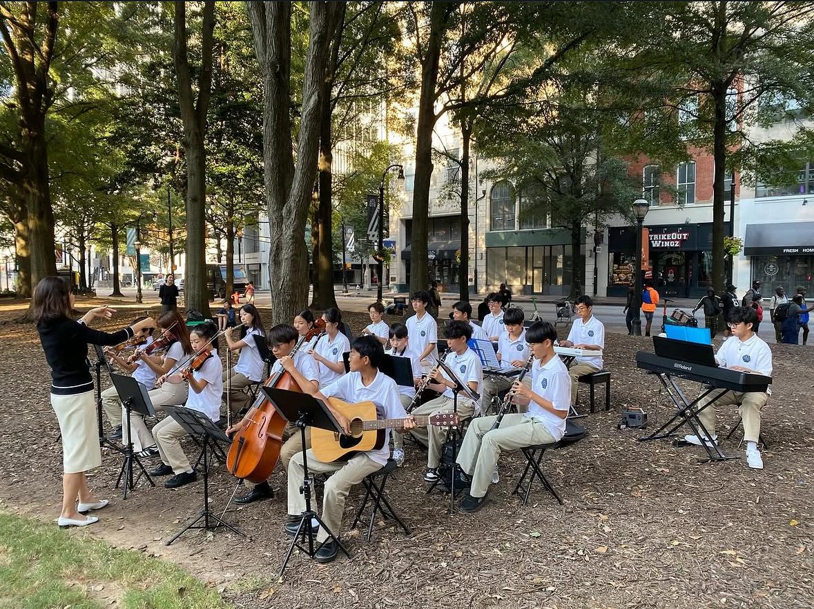 GYCO student orchestra performing outdoors in a downtown Atlanta park, with strings, clarinets, guitar, cello, and keyboards led by a conductor