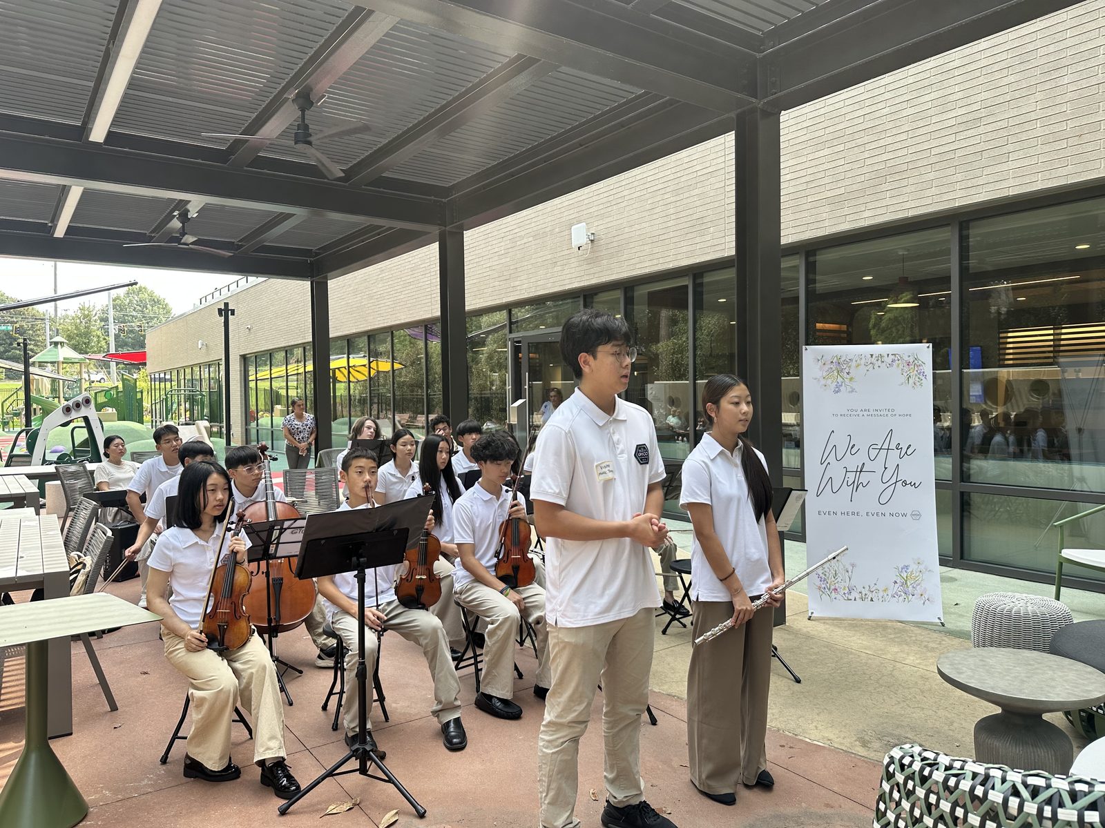 Two students introducing the ensemble's performance beside a We Are With You banner on the Ronald McDonald House Atlanta patio