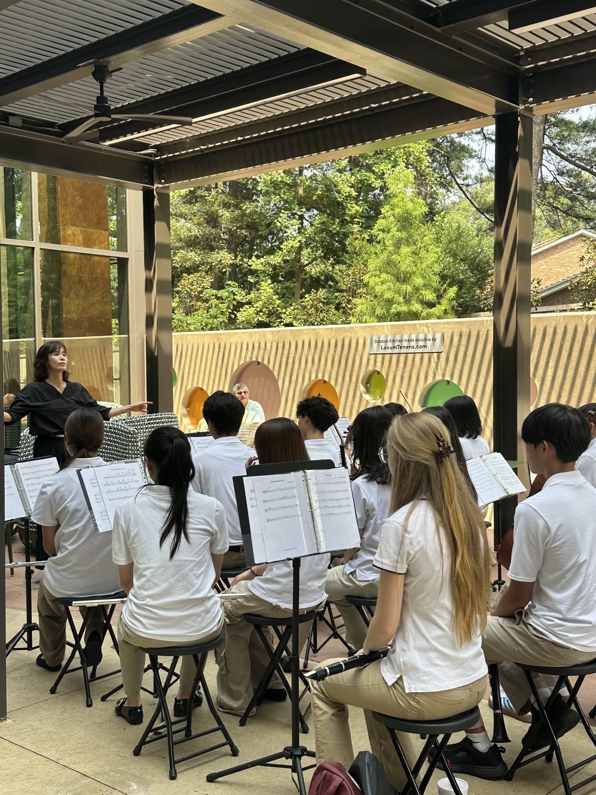 GYCO student ensemble seated with music stands under the outdoor pavilion at Ronald McDonald House Atlanta, following their conductor during a performance
